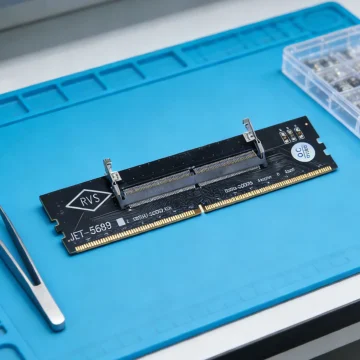 Black PCIe expansion card with gold edge connector and metal retention clips on a blue anti-static mat, labeled RVS, beside a small parts organizer box.