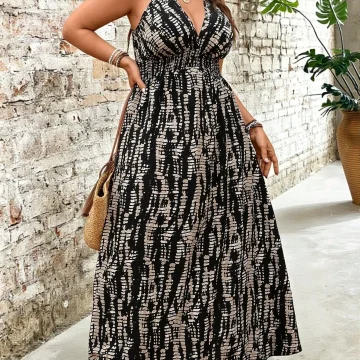 Woman in a black-and-white patterned maxi dress with a straw hat, sandals, and layered jewelry standing beside a stone wall.