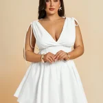Plus Size VNeck Drawstring Puffy Mini Dress 4 | PricZone Young woman in a white V neck dress with tied shoulders standing against a peach backdrop hands gently clasped wearing gold bracelets and star shaped earrings | PricZone