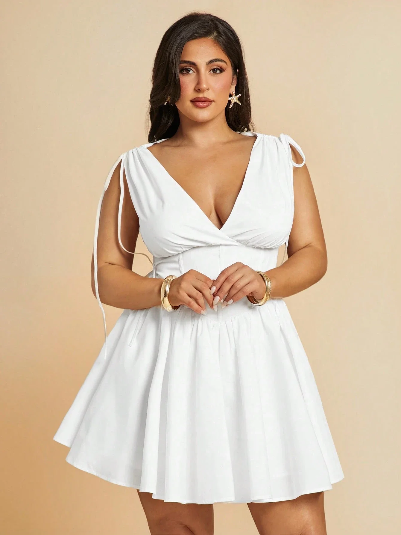 Plus Size VNeck Drawstring Puffy Mini Dress 4 | PricZone Young woman in a white V neck dress with tied shoulders standing against a peach backdrop hands gently clasped wearing gold bracelets and star shaped earrings | PricZone