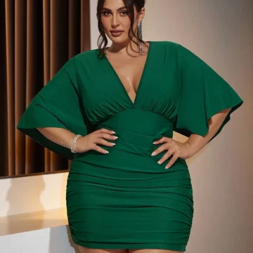 Woman in a emerald green, knee-length dress with a deep V-neck posing with hands on hips in an indoor setting.