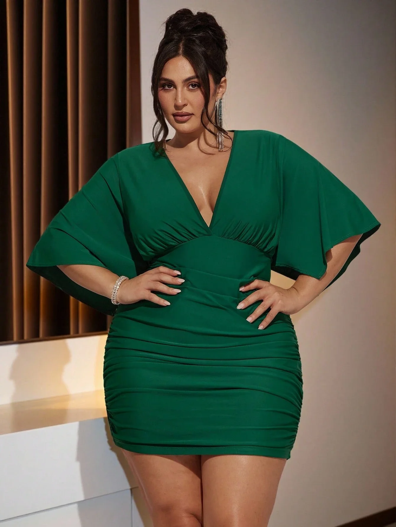Plus Size Women VNeck Ruched Mini Green Party Dress 2 | PricZone Woman in a emerald green knee length dress with a deep V neck posing with hands on hips in an indoor setting | PricZone