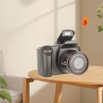 Digital camera with a flip out screen resting on a round wooden table in a softly lit living room | PricZone