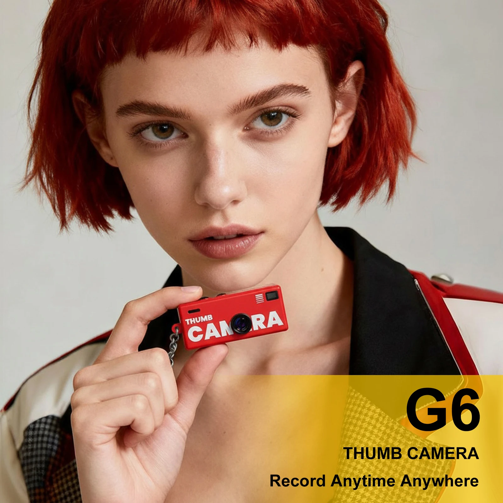 Close-up of a woman with short red hair holding a red mini camera labeled 'THUMB CAMERA' against a neutral backdrop.