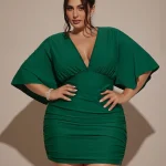Plus Size Women VNeck Ruched Mini Green Party Dress 5 | PricZone Woman in a emerald green ruched dress with a deep V neck posing with hands on hips in a studio setting wearing statement earrings and a bracelet | PricZone