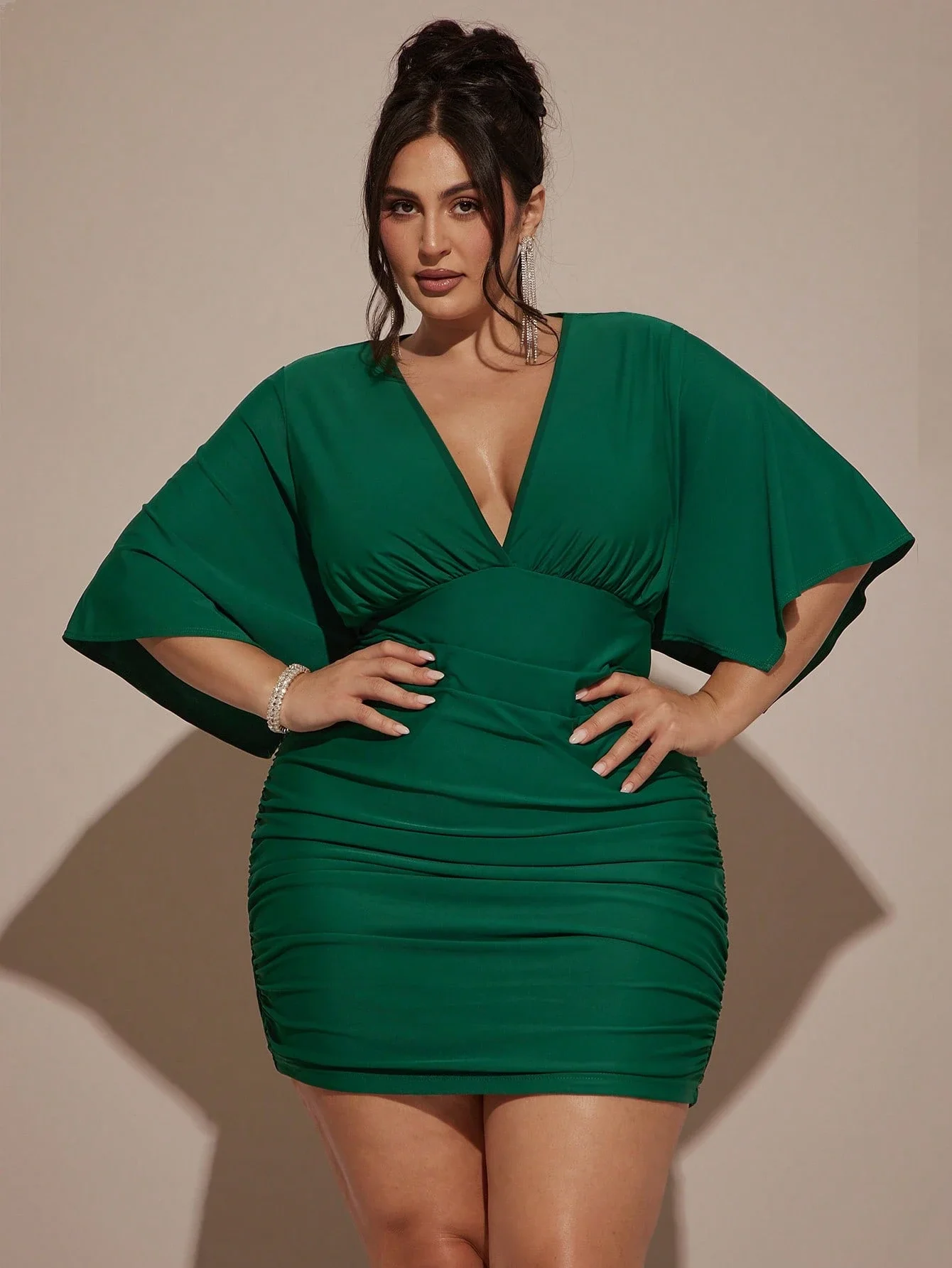 Plus Size Women VNeck Ruched Mini Green Party Dress 5 | PricZone Woman in a emerald green ruched dress with a deep V neck posing with hands on hips in a studio setting wearing statement earrings and a bracelet | PricZone