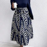 Plus Size Round Neck Long Sleeve Chevron Print Midi Dress 4 | PricZone Woman standing against a white wall wearing a navy top with puff sleeves and a navy and cream zigzag maxi skirt holding a white takeaway coffee cup | PricZone
