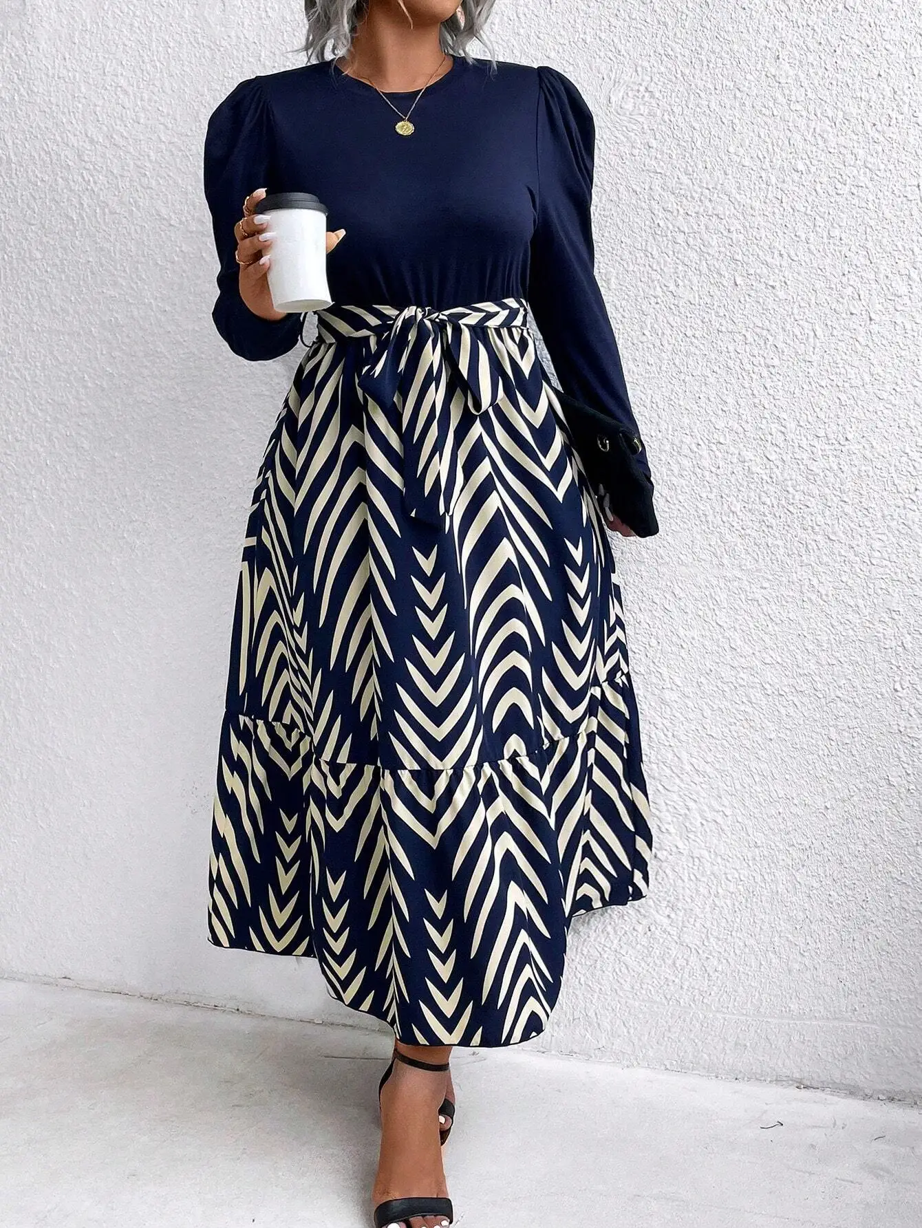 Plus Size Round Neck Long Sleeve Chevron Print Midi Dress 4 | PricZone Woman standing against a white wall wearing a navy top with puff sleeves and a navy and cream zigzag maxi skirt holding a white takeaway coffee cup | PricZone