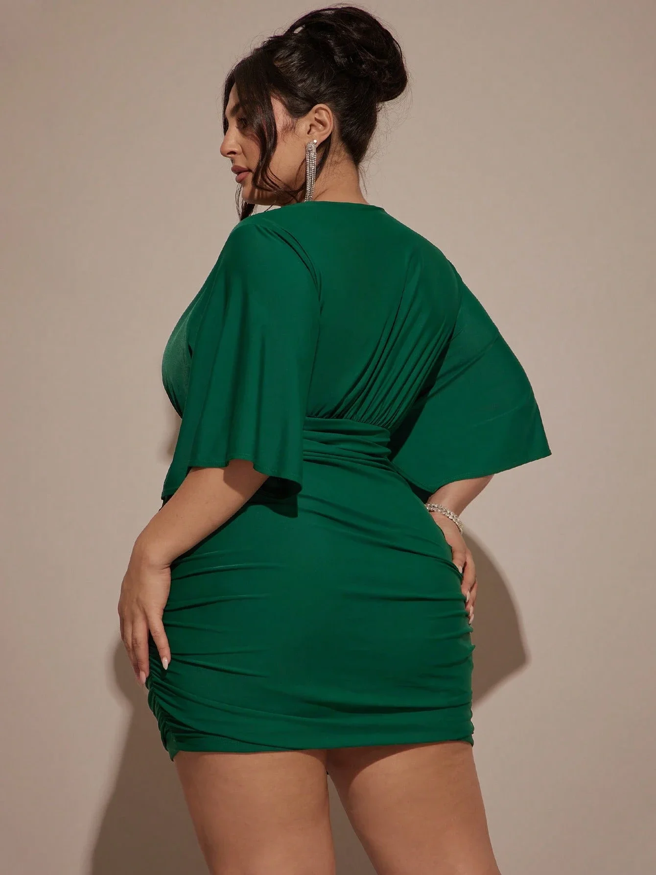 Plus Size Women VNeck Ruched Mini Green Party Dress 1 | PricZone Side view of a woman wearing a fitted emerald green dress with ruched detailing and draped sleeves her dark hair in an updo and wearing long earrings | PricZone
