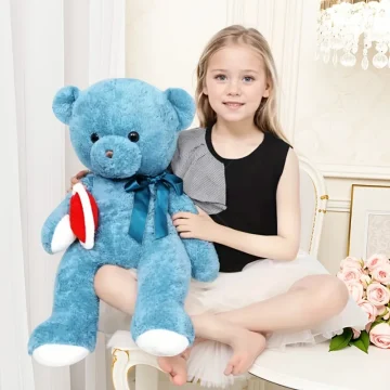 Young girl sits on a white chair, smiling, with a large blue teddy bear wearing a blue ribbon beside her.