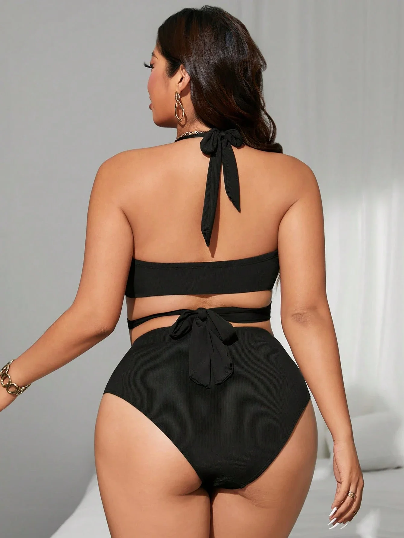 Black Plus Size Halter One Piece Swimsuit with Side Cutouts 2 | PricZone Woman wearing a black halter bikini from the back tie knot at neck and waist with gold jewelry visible on her left wrist and ear | PricZone