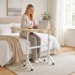 Small Standing Desk Adjustable Height with Wheels 5 | PricZone Woman seated on a bed typing on a laptop placed on a small portable table with a beige knit blanket and mug nearby in a bright cozy room | PricZone