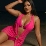 Woman in a bright pink halter swimsuit posing on a dimly lit beach at night with one arm resting above her head | PricZone