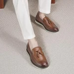 Brown leather tassel loafers worn with white pants shown from a top down angle on a beige carpeted floor | PricZone