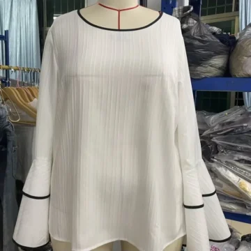 White pleated blouse on a mannequin, featuring black trim at the round neckline and layered bell sleeves with black edging, in a clothing store.
