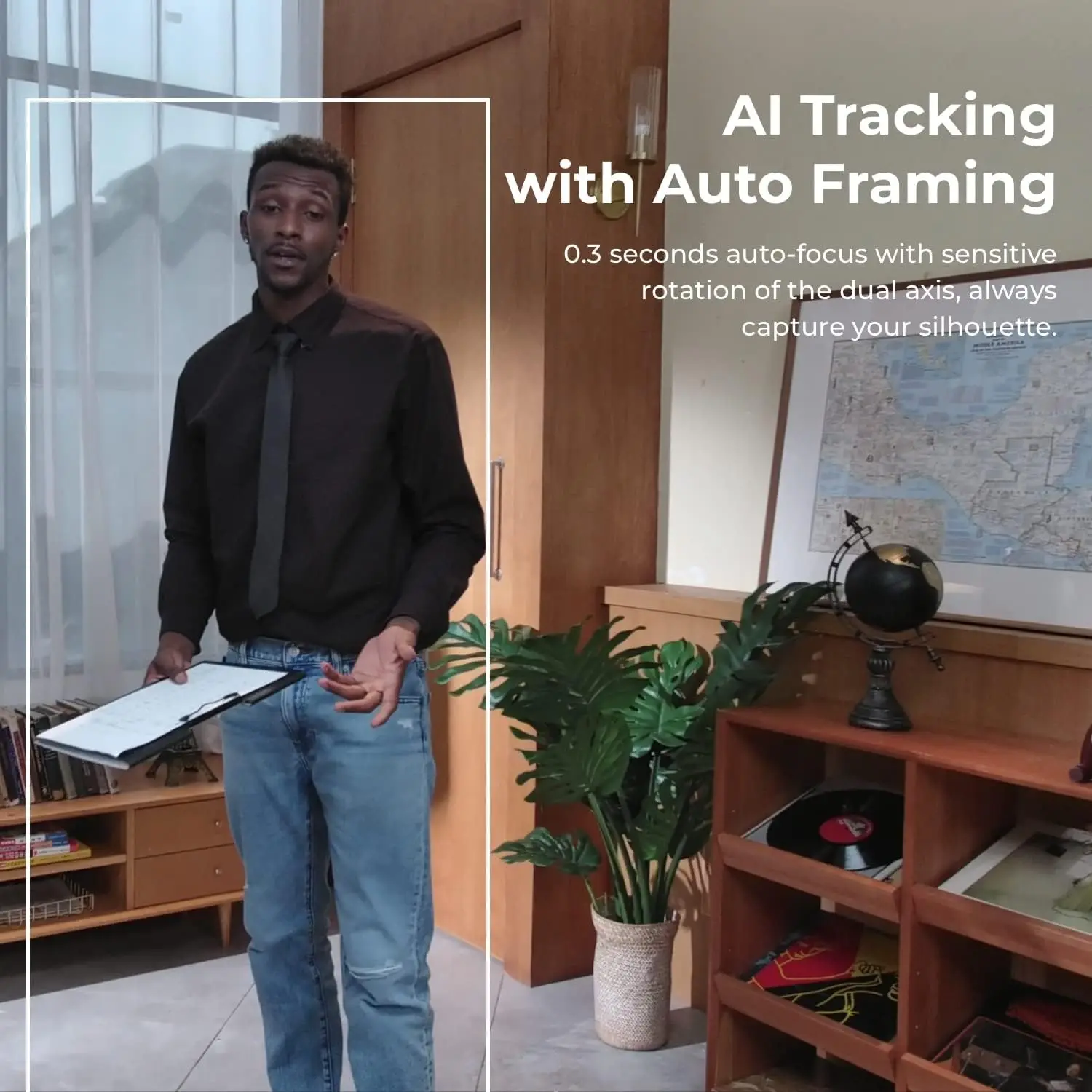 Man in a black shirt and jeans speaks, holding a clipboard in an office with a world map and globe on a shelf; white framing overlay visible for AI tracking.