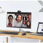 2K Qhd Webcam with Sony Sensor and Noise Reduction Mic 4 | PricZone Desk setup with a monitor displaying a six person video conference a laptop shows a chat app on a wooden desk | PricZone