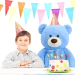 Boy in a striped shirt wearing a party hat beside a large blue teddy bear at a birthday party with colorful bunting and a decorated cake | PricZone