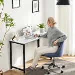 Woman in a gray knit sweater sits at a white desk using a laptop showing a video site in a bright home office with plants and framed art nearby | PricZone