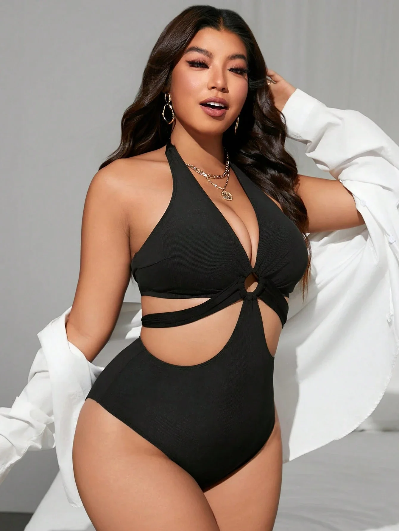 Black Plus Size Halter One Piece Swimsuit with Side Cutouts 3 | PricZone Woman in a black one piece swimsuit with cutouts gold jewelry and a white shirt draped over her arms posing against a light background | PricZone