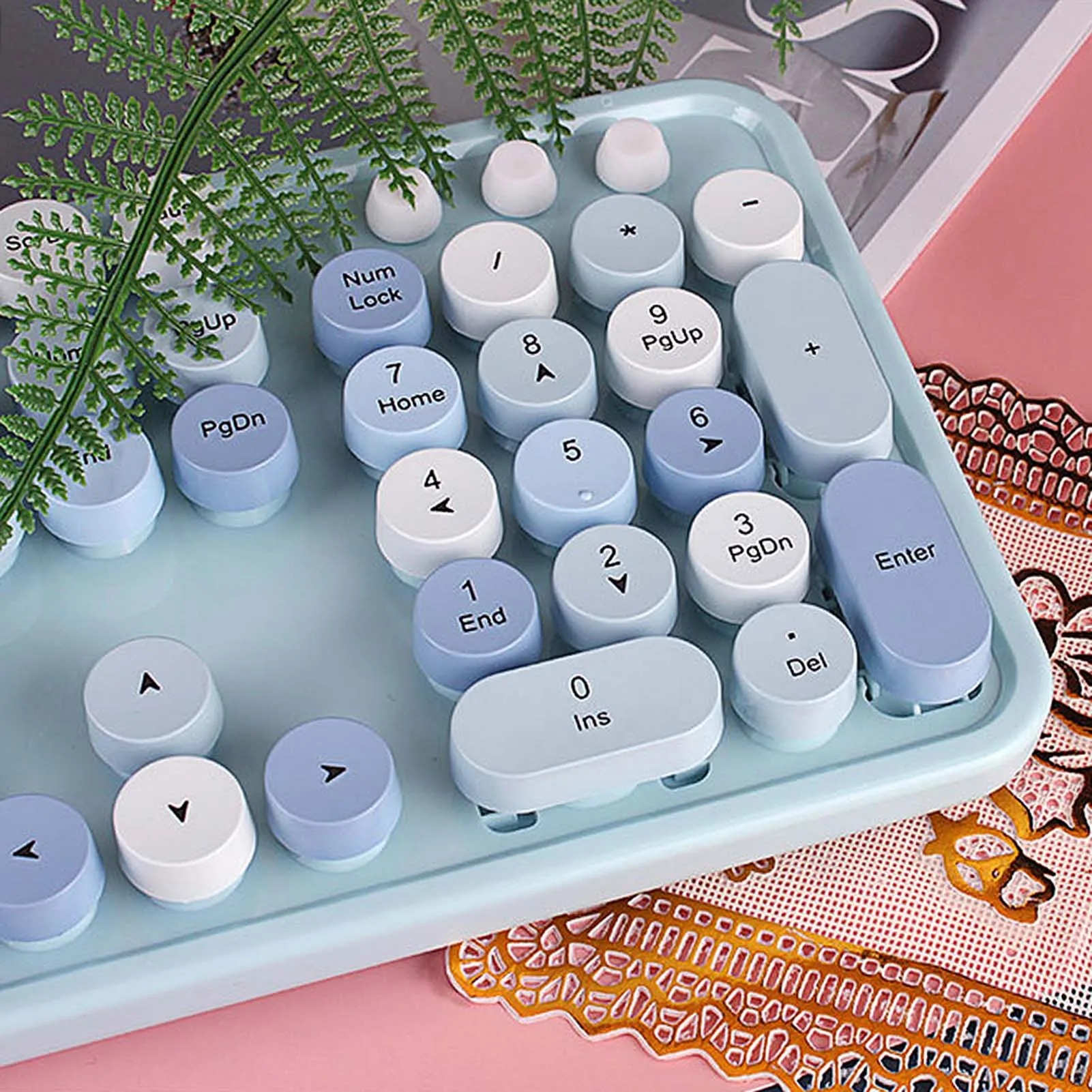 Pastel blue retro calculator with round, white and blue keys, a fern draped over the top, placed on a pink patterned surface.