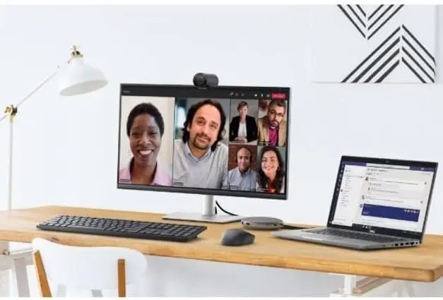 Desk setup with a six-person video call on a monitor; laptop, keyboard, and desk lamp on a wooden desk.