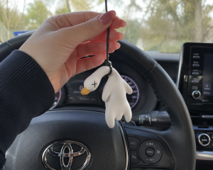 Hand holding a white duck shaped pendant with a black cord hanging from a car steering wheel | PricZone