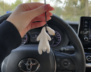 Hand suspends a white blob shaped plush figure on a black cord inside a car with the steering wheel and dashboard in the background | PricZone