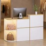 Modern boutique checkout counter white and wood desk with a computer monitor open shelves showing a beige purse and a brown handbag and a small potted plant on top clothing racks visible in the background | PricZone