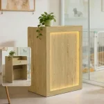Modern Reception Desk with Lockable Drawer 15Mm Mdf 1 | PricZone Tall light wood cabinet with a warm backlit inset panel and a potted plant on top placed by a glass door in a bright room | PricZone